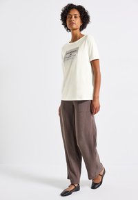 T-shirt blanche à manches courtes avec impression graphique, associée à un pantalon marron ample et des chaussures plates noires. Tissu lisse avec une texture subtile.