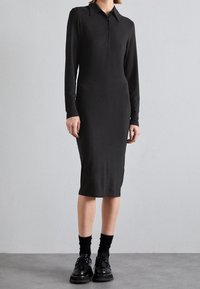 Schwarzes, geripptes Midikleid mit langen Ärmeln und spitzem Kragen, ausgestattet mit einer Knopfleiste. Kombiniert mit schwarzen Socken und Schuhen.