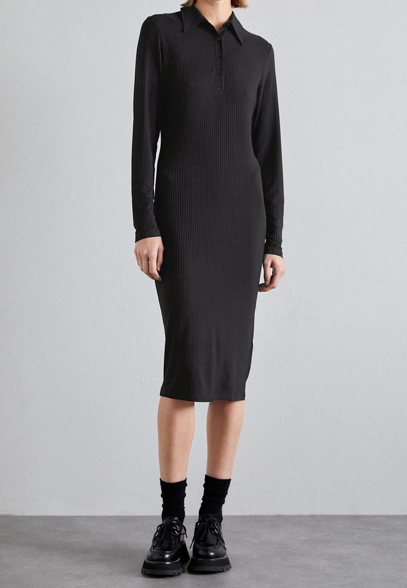 Schwarzes, geripptes Midikleid mit langen Ärmeln und spitzem Kragen, ausgestattet mit einer Knopfleiste. Kombiniert mit schwarzen Socken und Schuhen.
