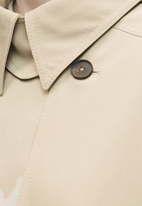 Collet et épaules d'un trench-coat beige avec un bouton marron foncé visible et des détails de couture soignés.