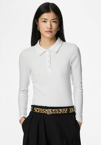 Biała koszulka polo z długim rękawem, z prążkowaną strukturą i kołnierzem, zapinana na guziki, zestawiona z czarnymi spodniami i paskiem w panterkę.