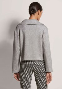 Graue, taillierte Jacke mit Reverskragen und langen Ärmeln, aus weichem Stoff gefertigt. Kombiniert mit gemusterten, hochgeschnittenen Hosen im geometrischen Design.