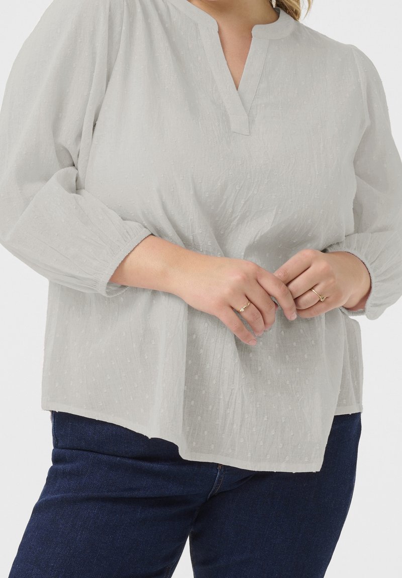 Femme portant un chemisier texturé gris clair à manches longues avec une encolure fendue et un jean bleu foncé, les mains délicatement jointes à la taille.