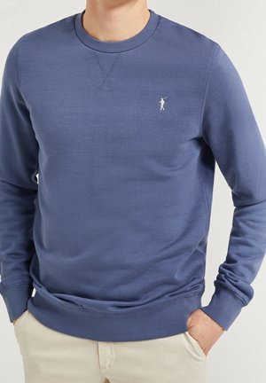 Mężczyzna ubrany w niebieską bluzę z okrągłym dekoltem z małym, białym haftowanym logo golfisty na piersi, w połączeniu z beżowymi spodniami.