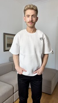 White textured short-sleeve shirt with a round neck and logo on the left chest, paired with black pants. Neutral indoor background.