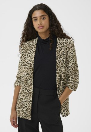 Žena s dlhými kučeravými vlasmi má na sebe sako s leopardím vzorom cez čierny rolák a čierne nohavice, stojí so jednou rukou v vrecku.