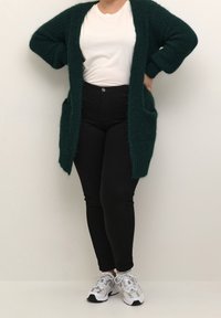 Lang, åben-front cardigan i mørkegrøn, lavet af et tekstureret, fluffy materiale; har to forlommer. Båret over en hvid t-shirt og sorte jeans.