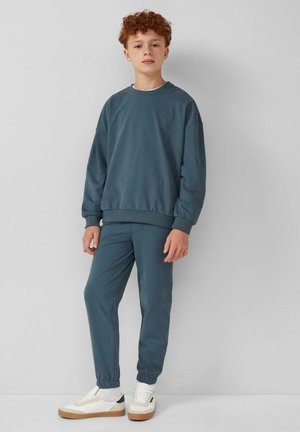 Donkerblauwe sweatshirt met een ontspannen pasvorm en geribbelde manchetten, gecombineerd met bijpassende taps toelopende broek, gedragen met witte sneakers en een lichtgekleurde shirt.