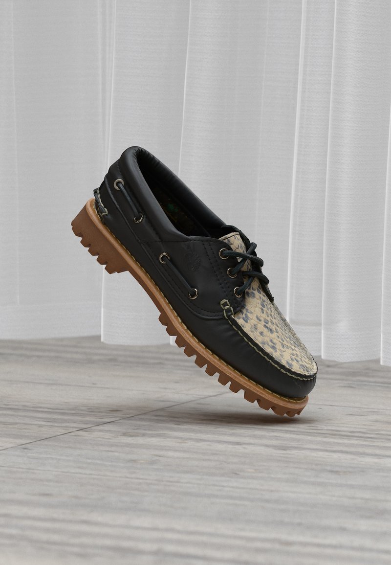 Μπεζ και μαύρο παπούτσι τύπου boat με σχέδιο λεοπάρδαλης, σκούρες κορδόνια και χοντρή καφέ πέλμα από καουτσούκ, επάνω σε φωτεινό ξύλινο πάτωμα μπροστά από λευκή κουρτίνα.