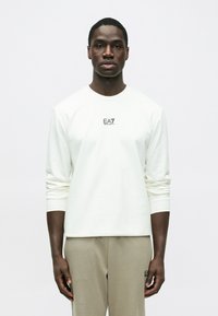 Tricou alb cu mânecă lungă, decolteu rotund, textură moale și logo negru EA7 pe piept. Asortat cu pantaloni bej deschis.