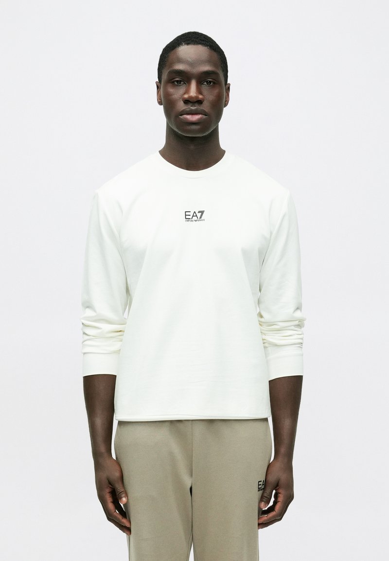 Tricou alb cu mânecă lungă, decolteu rotund, textură moale și logo negru EA7 pe piept. Asortat cu pantaloni bej deschis.