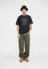 T-shirt grafico blu marina con logo a contrasto, pantaloni cargo verde oliva con tasche laterali, stivali marroni e cappello a secchiello nero.