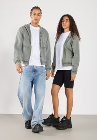 Felpe con cappuccio verde chiaro con zip e tasche frontali, t-shirt bianche, jeans blu e sneaker nere indossati da due modelli che si trovano su uno sfondo neutro.
