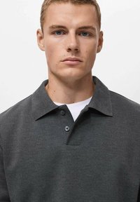 Dunkelgraues Poloshirt mit einem Kragenhals, drei Knöpfen und subtiler Textur. Über einem weißen Unterhemd getragen.