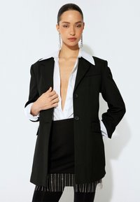 Blazer nero con colletto bianco a contrasto, design monopetto, due bottoni e tasche. Indossato sopra una camicetta bianca con scollatura a V profonda.