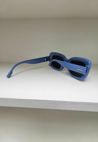 Lunettes de soleil bleues avec une monture carrée, fabriquées en plastique, dotées de verres teintés foncés et d'un logo sur la branche. Le design est épais et moderne.