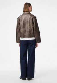 Veste en cuir marron avec un design court et des manches larges, associée à un jean large bleu foncé. Texture lisse avec peu de matériel.