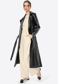 Lungo trench coat in pelle nera con vita cinturata, abbinato a pantaloni larghi crema e ballerine con fiocco in bianco e nero.
