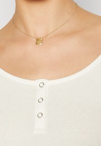 Kultainen kaulakoru, jossa neliömäinen, kuvioitu riipus, puettuna valkoisen ribbineuletopin päälle hopeanväristen nappikoristeiden kanssa.