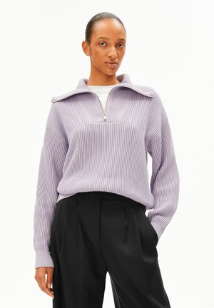 Personne portant un pull à fermeture éclair côtelé violet clair par-dessus un haut blanc et un pantalon plissé noir, debout avec une main dans la poche.