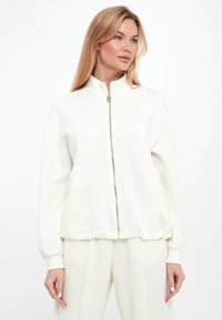 Giacca bianca con zip e colletto alto, realizzata in tessuto morbido. Presenta polsini e orlo a coste, e un design frontale liscio.