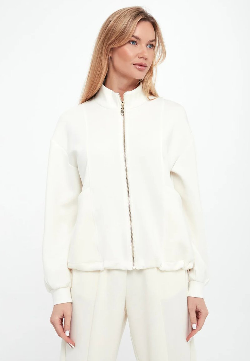 Giacca bianca con zip e colletto alto, realizzata in tessuto morbido. Presenta polsini e orlo a coste, e un design frontale liscio.