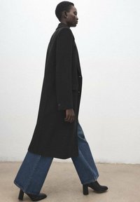 Długi czarny płaszcz o dopasowanym kroju, noszony na szerokich spodniach dżinsowych o prostych nogawkach. Charakteryzuje się podkreślonymi ramionami i czarnymi szpiczastymi szpilkami.