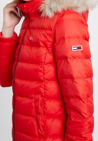 Veste rembourrée rouge avec une capuche doublée de fourrure, présentant des sections matelassées, une fermeture éclair sur le devant et un patch logo sur la manche gauche.