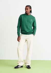 Green cotton sweatshirt, cream striped pants, black sneakers with white stripes. Model stands on a green floor against a white backdrop.