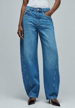 Persona vistiendo jeans azules de tiro alto y pierna ancha, combinados con tacones negros de punta puntiaguda, de pie con una mano en el bolsillo y la otra relajada.