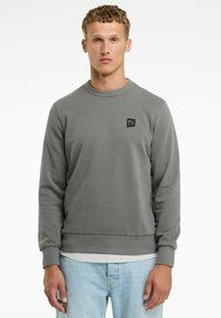 Felpa grigia con superficie texturizzata, polsini a coste e colletto a girocollo. Presenta un piccolo logo nero sul petto. Indossata sopra jeans leggeri blu.