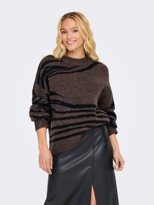 Donna bionda sorridente, indossa un maglione a righe marroni e nere con le maniche arrotolate e una gonna di pelle nera con spacco frontale.