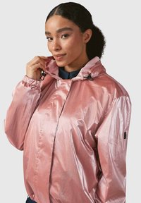 Könnyű, fényes rózsaszín dzseki, elülső cipzárral, kapucnival és elastikus mandzsettákkal. Lazább szabású és minimalista dizájnnal rendelkezik, húzózsinórokkal.