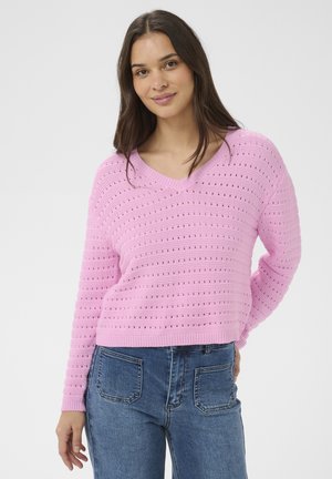 Mladá žena s dlhými hnedými vlasmi, oblečená v ružovom dierkovanom svetri s véčkovým výstrihom a modrých džínsach, pózujúca s jednou rukou za chrbtom.