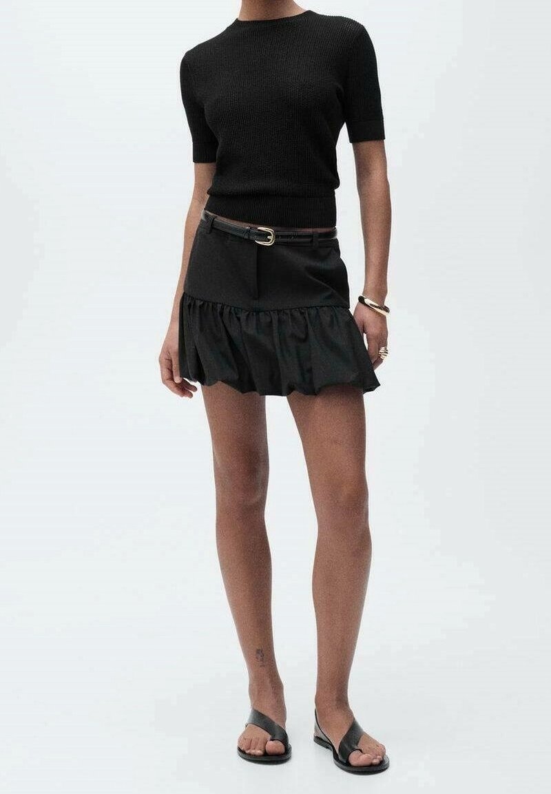 Femme portant un haut en maille noir à manches courtes, une mini-jupe noire à volants avec ceinture, des sandales noires, et un bracelet ainsi que des bagues en or.