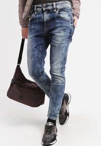 Man med urtvättade slim-fit blå jeans, svart och grå sneakers, hållande en mörkbrun axelväskan, med en hand i fickan.