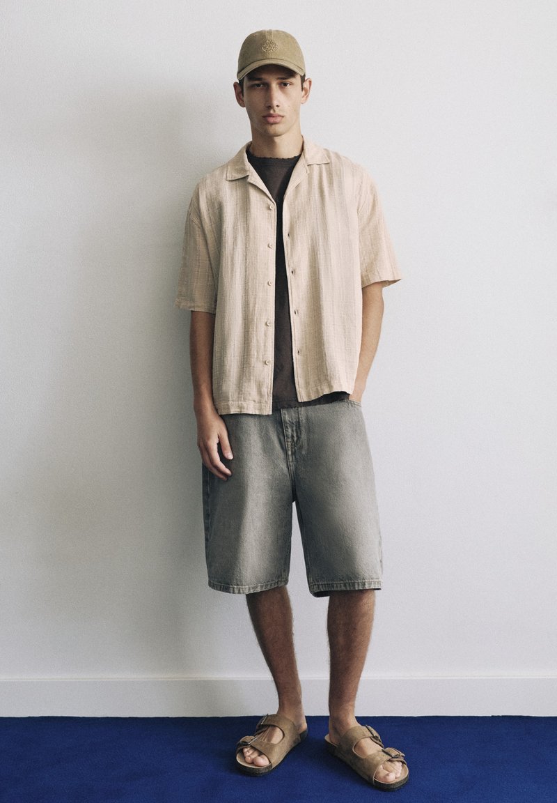 Jeune homme debout contre un mur uni, portant une chemise beige à manches courtes, un short gris, des sandales beige et une casquette de baseball marron.