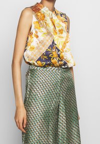 Blouse sans manches avec un mélange de motifs floraux jaunes, oranges et bleu marine, dotée d'un décolleté en trou de serrure et d'un tissu doux et texturé. Pantalon à motifs verts.