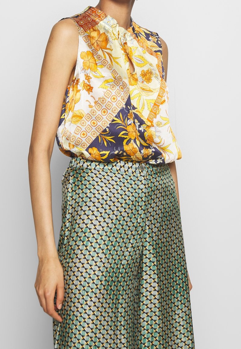 Blouse sans manches avec un mélange de motifs floraux jaunes, oranges et bleu marine, dotée d'un décolleté en trou de serrure et d'un tissu doux et texturé. Pantalon à motifs verts.