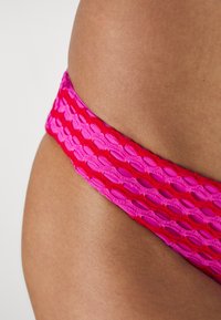 Costum de baie inferior roz și roșu, cu un design texturat și împletit, prezentând un model în diamant, punând accent pe culori vibrante și o bandă elastică.