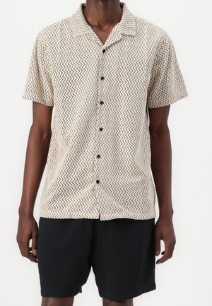 Mand iført en beige kortærmet skjorte med knapper og et zigzag-mønster samt sorte shorts, stående mod en ensfarvet hvid baggrund.