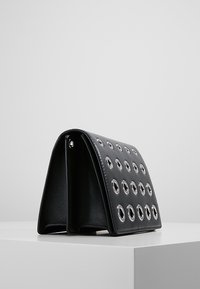 Bolso de mano negro de cuero con bordes redondeados, con múltiples detalles de ojales plateados en la parte frontal, interior texturizado y apoyado sobre una superficie plana.