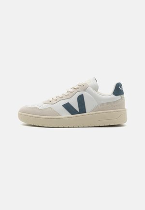 Baskets en cuir blanc avec des accents en suede gris, arborant un logo "V" bleu bien visible, une pointe ronde et une semelle en caoutchouc texturée.