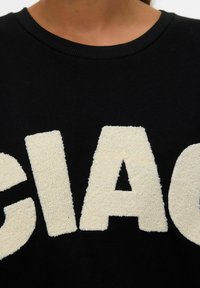 Zwarte sweatshirt met grote, crèmekleurige, donzige terrycloth letters die "GIAG" spellen. Ribbelneklijn en zachte textuur. Close-up weergave.