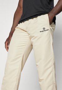 Beige treningsbukser med slim fit, laget av glatt stoff. Har en svart logo på venstre lår og dype lommer på siden.