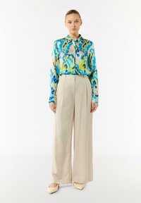 Kleurrijk zijden shirt met blauw, groen en geel abstract patroon, gecombineerd met hooggepunte beige wijde broek en lichtbeige schoenen.