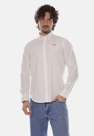 Uomo con capelli ricci che indossa una camicia bianca abbottonata con piccolo logo, jeans blu, in piedi con una mano in tasca contro uno sfondo semplice.