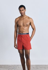 Κόκκινα μαγιό σορτς με κορδόνι και μπλε ναυτικό ζωνάρι στη μέση με λογότυπο της μάρκας· λεία υφή, μήκος έως το γόνατο και άνετη εφαρμογή.