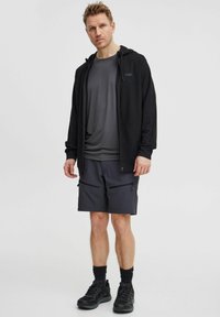 Svart hettegenser over en grå T-skjorte, sammen med mørke shorts med glidelåslommer. Sorte joggesko og svarte sokker fullfører antrekket.