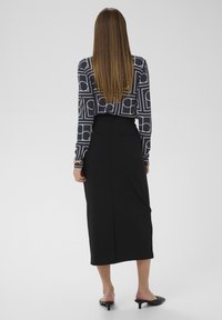 Falda negra de talle alto con una abertura trasera, combinada con una blusa azul marino con diseños geométricos blancos; modelo vista desde atrás.